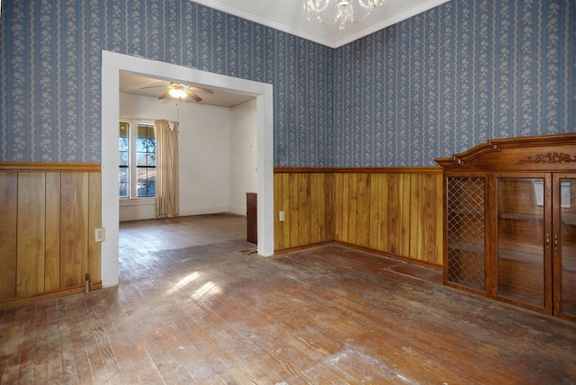 unfurnished room with a wainscoted wall, ceiling fan with notable chandelier, wallpapered walls, wood-type flooring, and crown molding