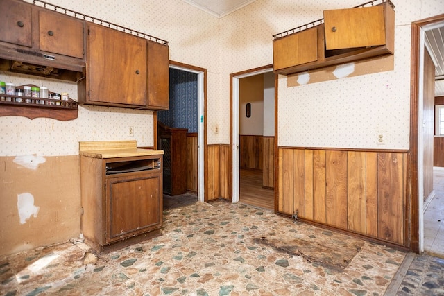 kitchen featuring a wainscoted wall, wood walls, brown cabinetry, and wallpapered walls