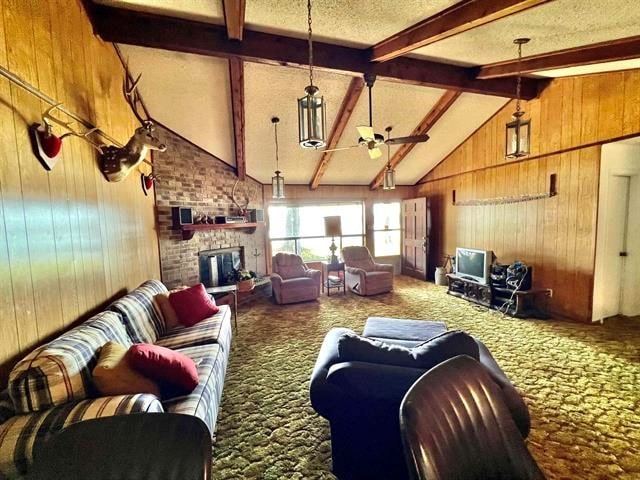 carpeted living room featuring a textured ceiling, ceiling fan, wooden walls, a fireplace, and vaulted ceiling with beams