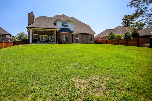 back of house featuring a ceiling fan, a chimney, a fenced backyard, a lawn, and brick siding