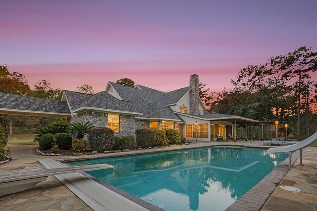 pool at dusk with a patio area, a diving board, and a water slide