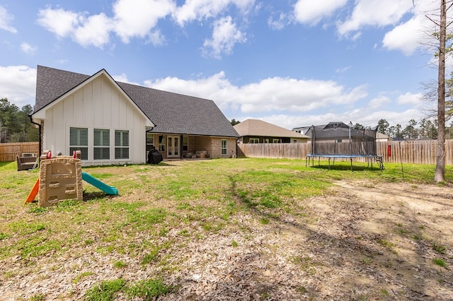 rear view of house featuring a fenced backyard, a shingled roof, a trampoline, a lawn, and board and batten siding