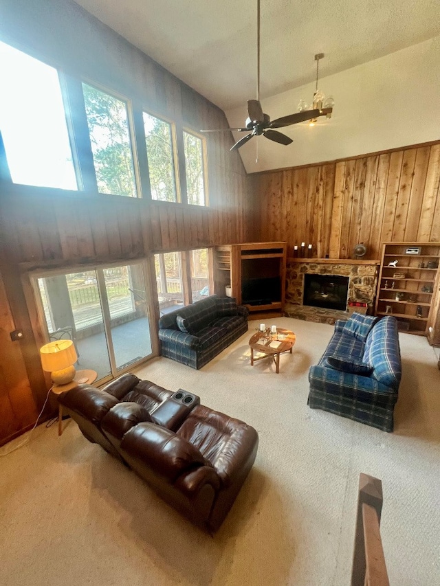 carpeted living room with a ceiling fan, a high ceiling, a stone fireplace, wood walls, and a wealth of natural light