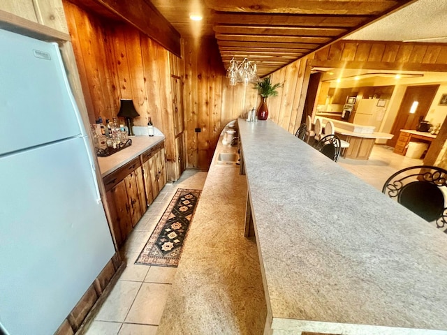 bar featuring light tile patterned flooring, beam ceiling, wood walls, and freestanding refrigerator