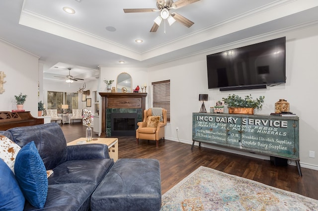 living room with crown molding, ceiling fan, a tray ceiling, a premium fireplace, and dark wood-type flooring