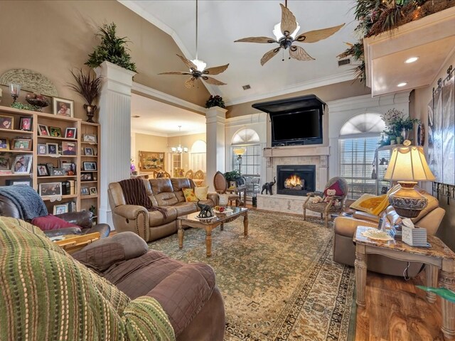 living room with a warm lit fireplace, ceiling fan with notable chandelier, wood finished floors, ornamental molding, and decorative columns