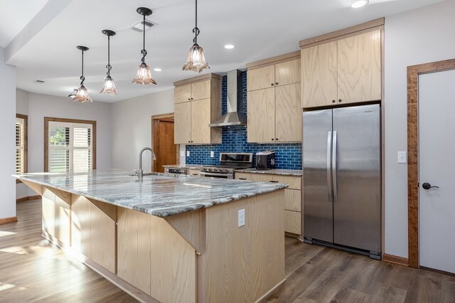 kitchen featuring wall chimney range hood, sink, an island with sink, light brown cabinetry, and appliances with stainless steel finishes