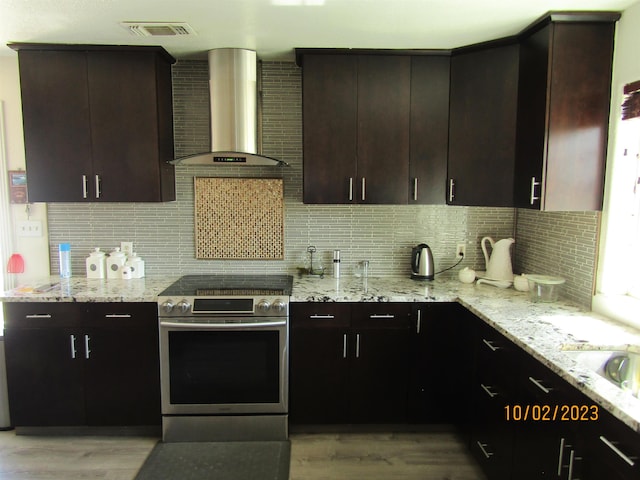 kitchen with stainless steel stove, wall chimney exhaust hood, light stone countertops, light hardwood / wood-style floors, and dark brown cabinetry