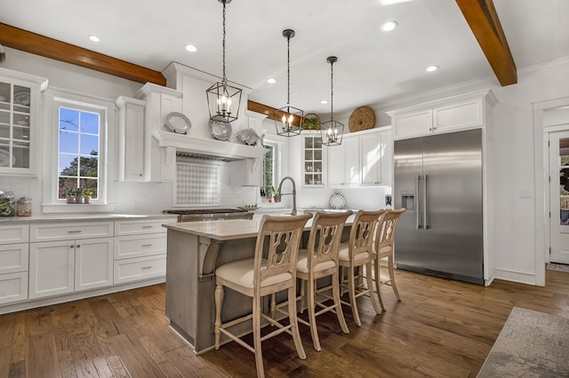 kitchen featuring backsplash, glass insert cabinets, a center island with sink, beam ceiling, and built in fridge