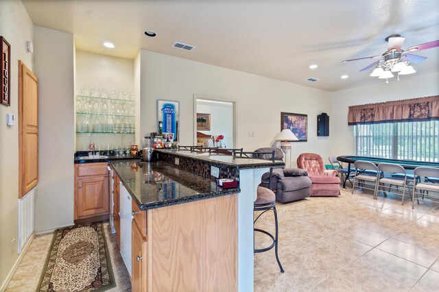 kitchen featuring ceiling fan, light brown cabinets, dark stone counters, a kitchen bar, and light tile patterned floors