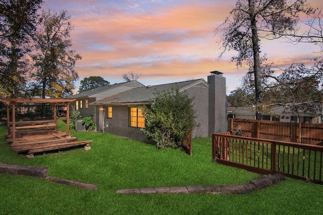 back of house at dusk with a lawn, a chimney, fence, a wooden deck, and brick siding