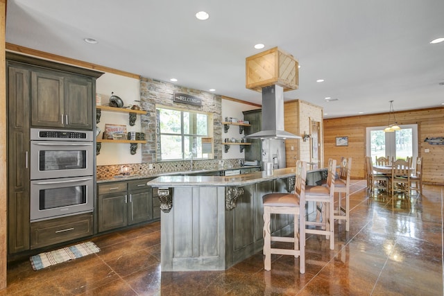 kitchen featuring island exhaust hood, wood walls, stainless steel double oven, and a healthy amount of sunlight