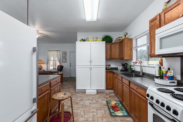 kitchen with dark countertops, a sink, brown cabinets, white appliances, and stone finish flooring
