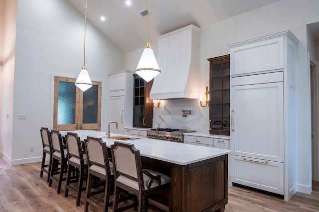 kitchen with premium range hood, an island with sink, backsplash, light wood-style floors, and a breakfast bar area