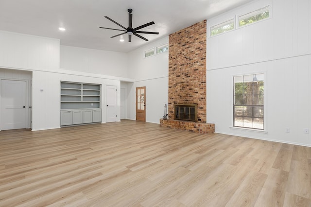 unfurnished living room featuring built in shelves, ceiling fan, a high ceiling, a brick fireplace, and light hardwood / wood-style flooring