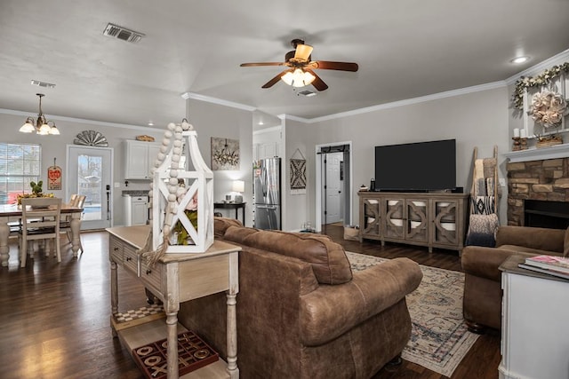 living room with visible vents, ornamental molding, ceiling fan with notable chandelier, a stone fireplace, and dark wood-style flooring