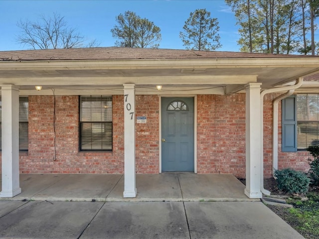 property entrance featuring brick siding, covered porch, and roof with shingles