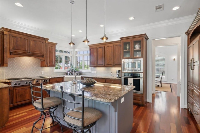 kitchen with light stone countertops, stainless steel appliances, crown molding, dark wood-type flooring, and an island with sink