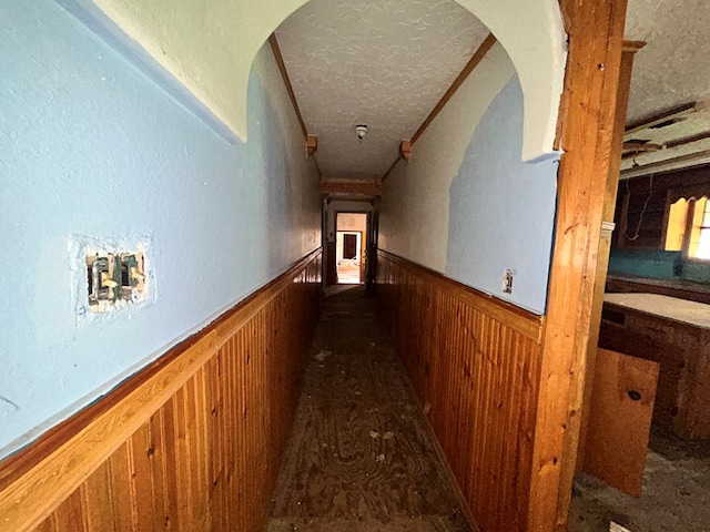 hall with arched walkways, wooden walls, a textured ceiling, and a wainscoted wall