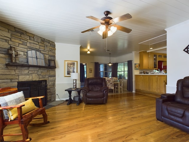 living room with a stone fireplace, crown molding, ceiling fan, and light wood-type flooring