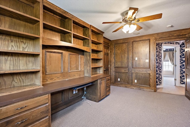 carpeted home office featuring wood walls, a textured ceiling, ceiling fan, and built in desk