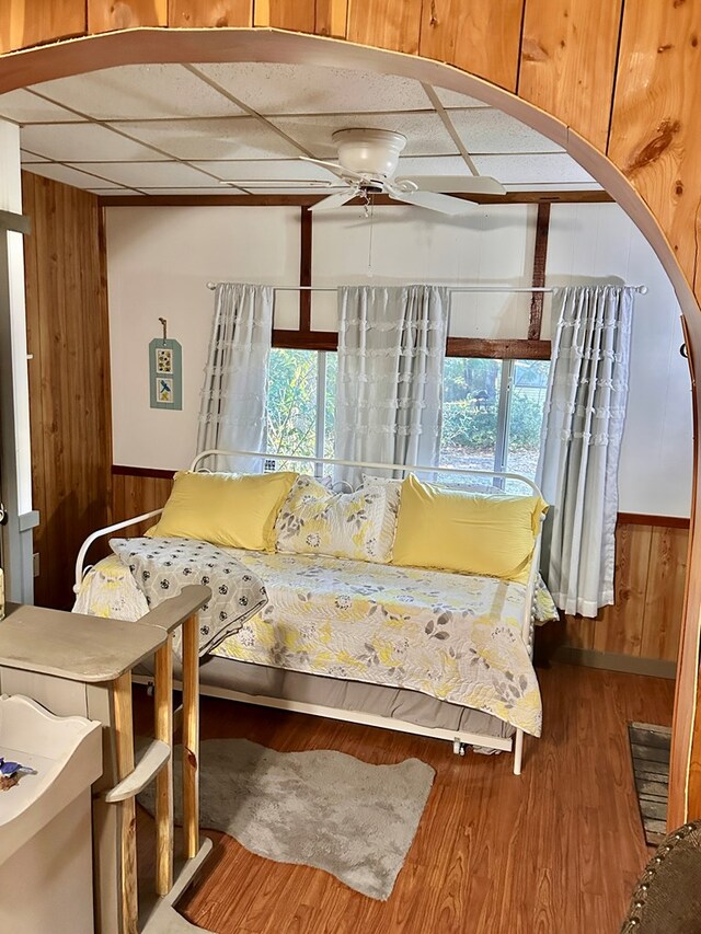 bedroom featuring ceiling fan, wooden walls, wood-type flooring, and a drop ceiling