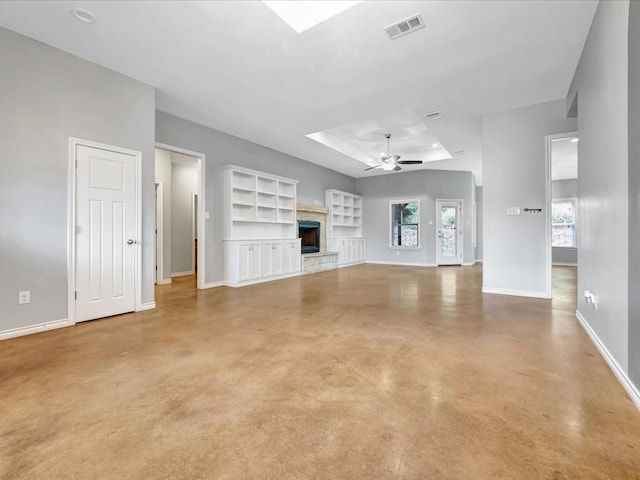 unfurnished living room with a raised ceiling, built in shelves, ceiling fan, and a stone fireplace