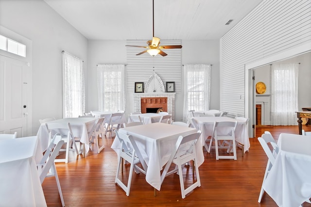 dining room with ceiling fan, wood-type flooring, a towering ceiling, and a brick fireplace