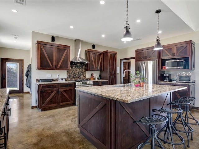 kitchen with appliances with stainless steel finishes, hanging light fixtures, a kitchen island with sink, wall chimney range hood, and a sink