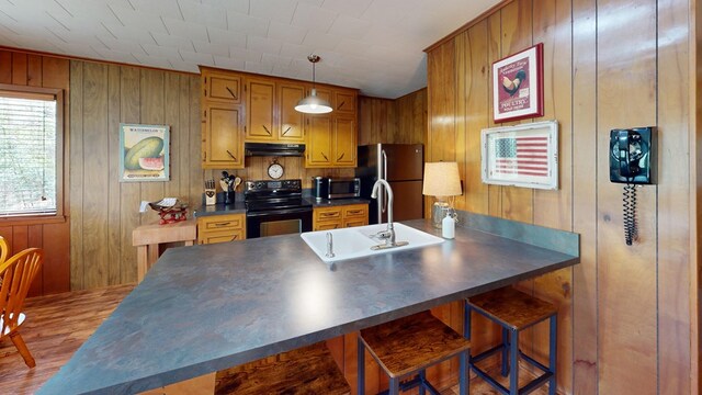 kitchen featuring wood walls, dark countertops, under cabinet range hood, a sink, and black appliances