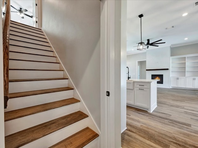 stairway featuring wood finished floors, visible vents, recessed lighting, a warm lit fireplace, and ceiling fan