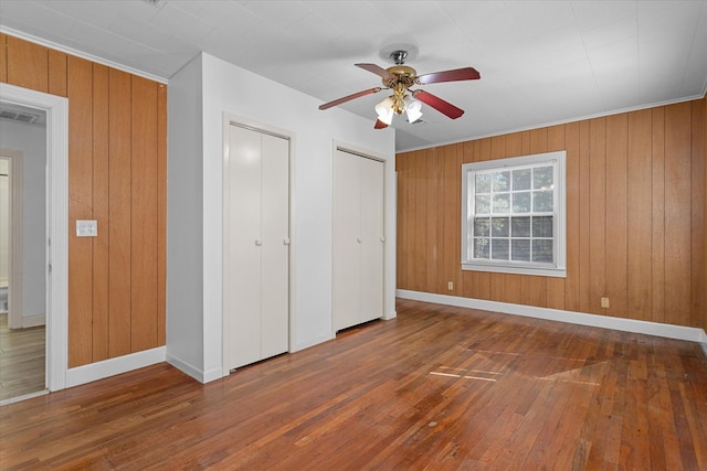 unfurnished bedroom with wood walls, two closets, ceiling fan, ornamental molding, and dark hardwood / wood-style flooring