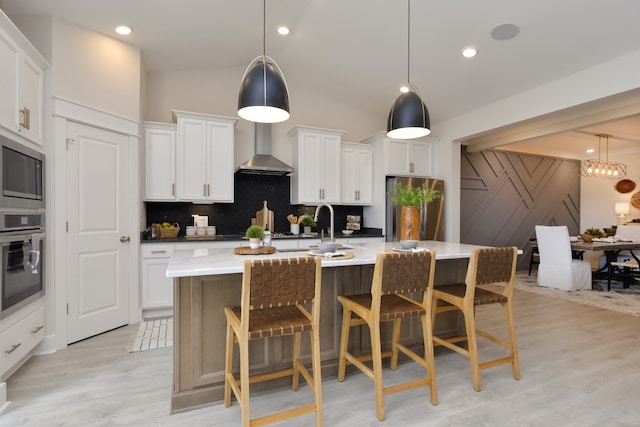 kitchen featuring appliances with stainless steel finishes, an island with sink, a notable chandelier, light hardwood / wood-style flooring, and wall chimney exhaust hood