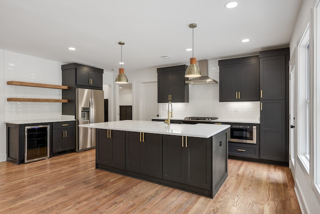 kitchen featuring decorative light fixtures, wine cooler, light hardwood / wood-style flooring, wall chimney exhaust hood, and a kitchen island with sink