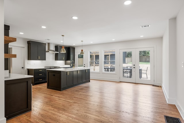 kitchen featuring light hardwood / wood-style floors, an island with sink, wall chimney exhaust hood, french doors, and pendant lighting