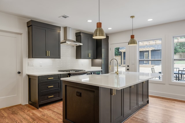 kitchen featuring an island with sink, pendant lighting, backsplash, light hardwood / wood-style floors, and wall chimney range hood