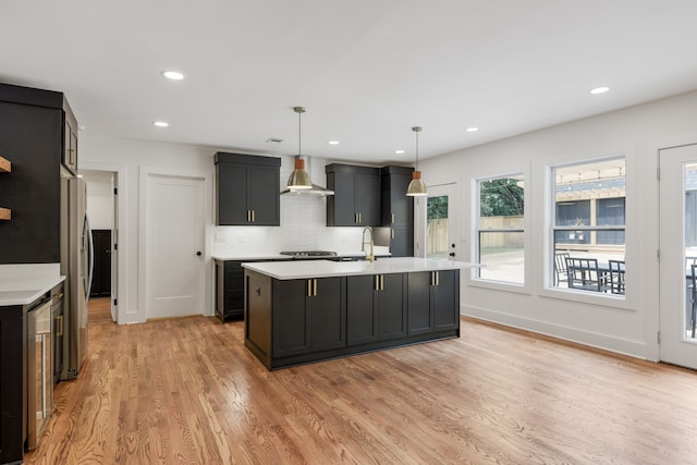 kitchen with wall chimney range hood, hanging light fixtures, a kitchen island with sink, and light wood-type flooring