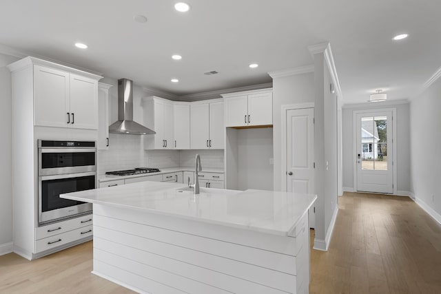 kitchen featuring a kitchen island with sink, appliances with stainless steel finishes, white cabinets, wall chimney range hood, and ornamental molding