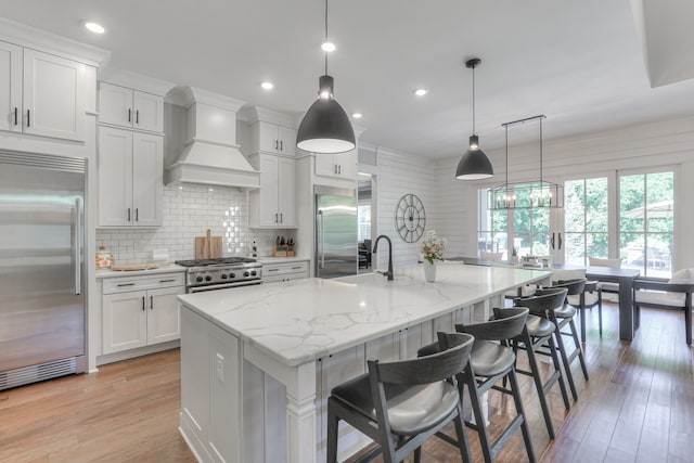 kitchen featuring high quality appliances, light hardwood / wood-style floors, custom exhaust hood, hanging light fixtures, and a kitchen island with sink