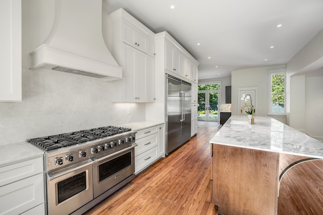 kitchen with premium range hood, light hardwood / wood-style floors, light stone countertops, high end appliances, and a kitchen island with sink