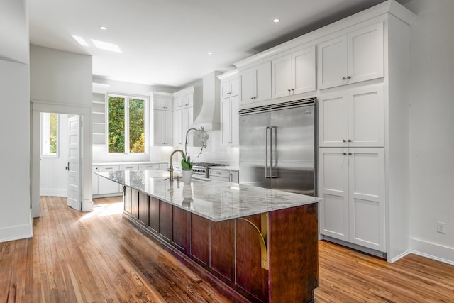 kitchen with hardwood / wood-style flooring, custom exhaust hood, white cabinets, stainless steel built in refrigerator, and light stone counters