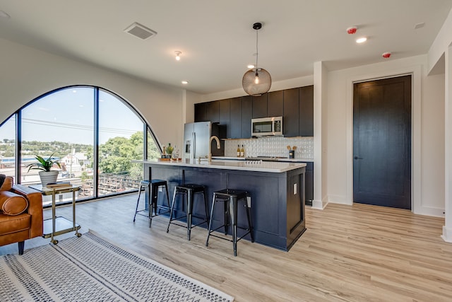 kitchen featuring appliances with stainless steel finishes, pendant lighting, backsplash, light hardwood / wood-style flooring, and a kitchen island with sink