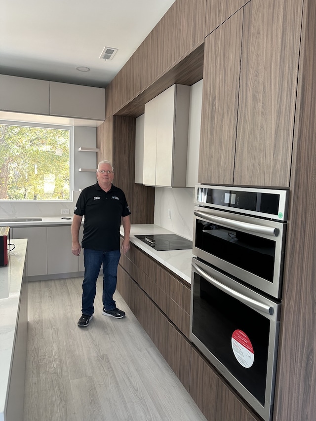 kitchen featuring black electric stovetop, stainless steel double oven, light stone counters, and light wood-type flooring