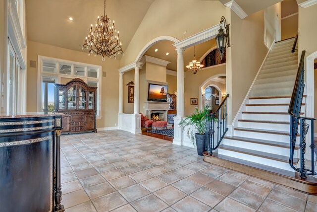 foyer featuring a warm lit fireplace, decorative columns, a chandelier, stairs, and high vaulted ceiling