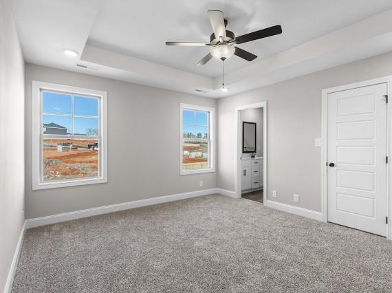 unfurnished bedroom with ceiling fan, ensuite bathroom, a raised ceiling, and carpet flooring