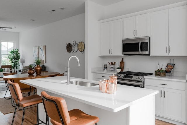 kitchen with ceiling fan, a kitchen island with sink, light hardwood / wood-style floors, stainless steel appliances, and white cabinets