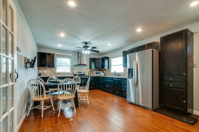 kitchen featuring backsplash, ceiling fan, light hardwood / wood-style floors, stainless steel appliances, and wall chimney exhaust hood