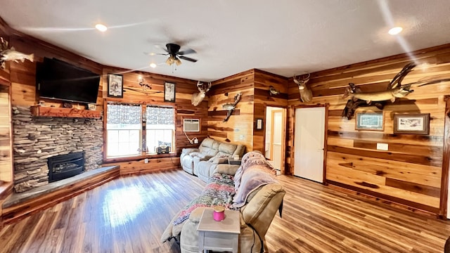 living room featuring ceiling fan, a wood stove, a stone fireplace, wooden walls, and hardwood / wood-style flooring