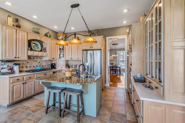 kitchen featuring light brown cabinets, backsplash, decorative light fixtures, a center island with sink, and appliances with stainless steel finishes