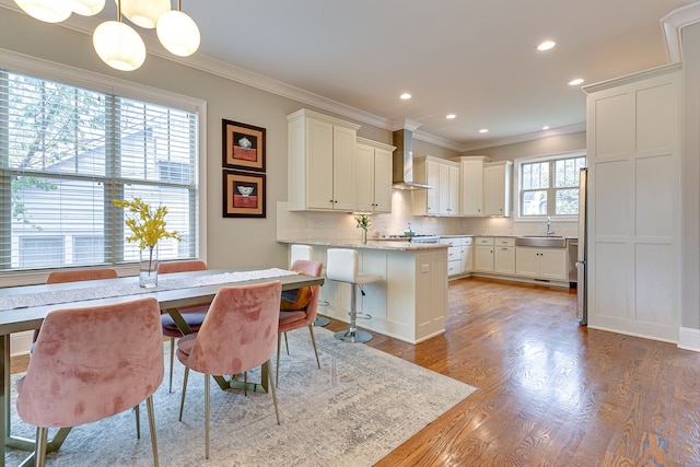 kitchen featuring dark wood-type flooring, a kitchen bar, pendant lighting, wall chimney exhaust hood, and ornamental molding